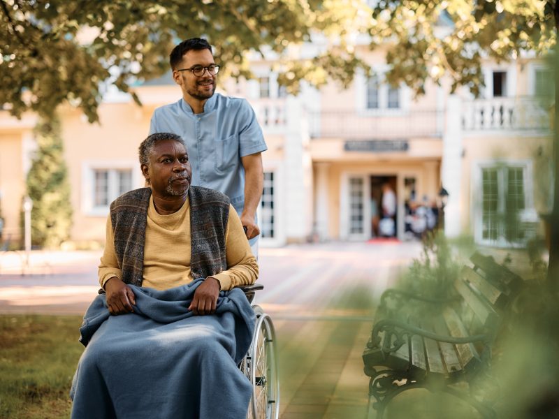 Young healthcare worker pushing senior black man in wheelchair through nursing home's park.